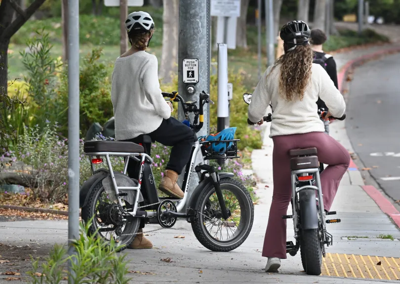 Two people sitting on stationary electronic bikes at a crosswalk entrance