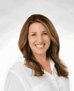 A woman with long, light brown hair and a bright smile wears a white shirt and is standing against a plain, light background.